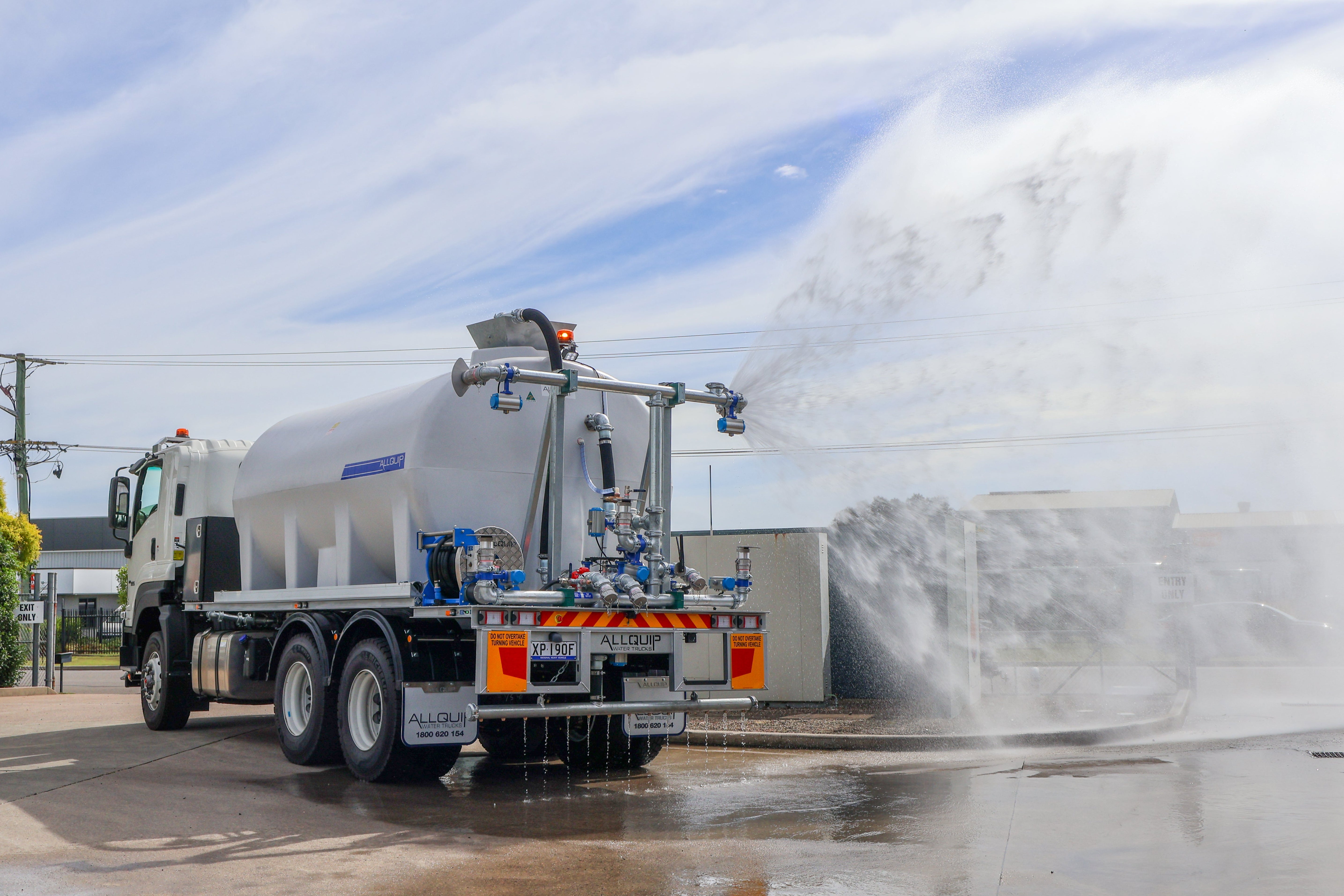 A white water truck spraying water outdoors under a blue sky, with a large water mist spraying to the right.